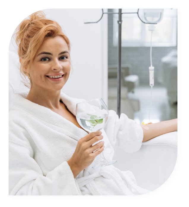 Woman in a white robe relaxing with a drink while receiving an IV drip in a modern, serene setting.